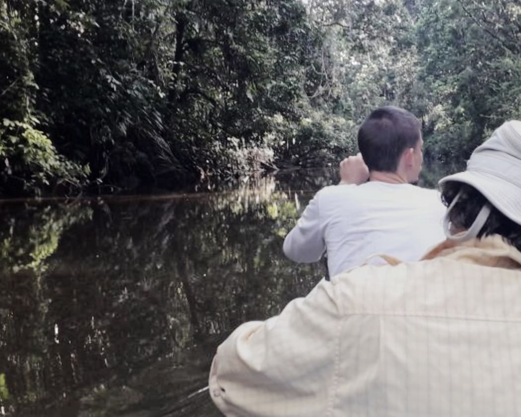 A man and a boy paddle a canoe up a dark river reflecting the jungle on the shore