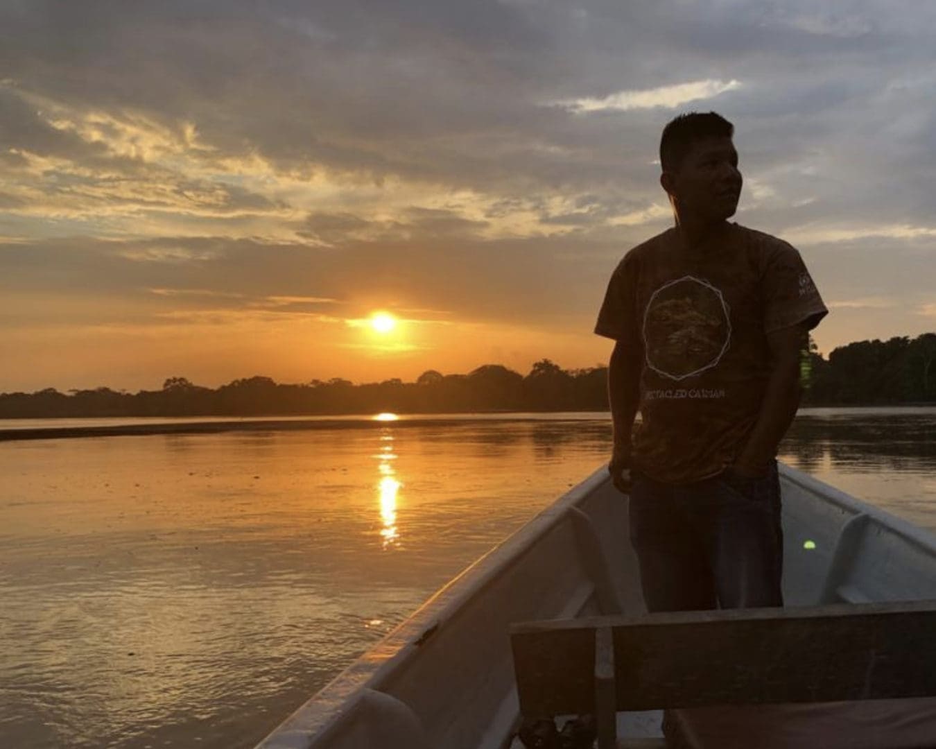 Man stands in bow of canoe with setting sun reflected in water in the background