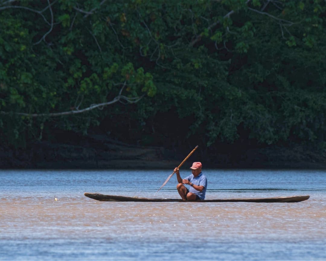 A man paddles a traditional wooden canoe on the deep blue, gray river backed by dense jungle foliage